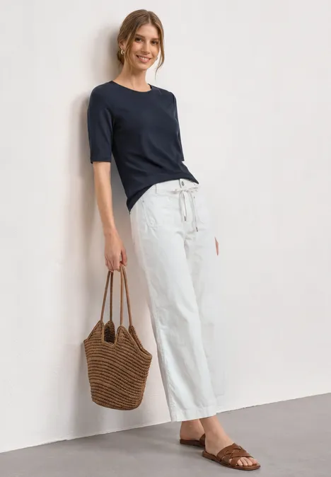 Vrouw leunt tegen muur, draagt donkerblauw shirt, wijde witte broek, bruine sandalen en gevlochten tas.
