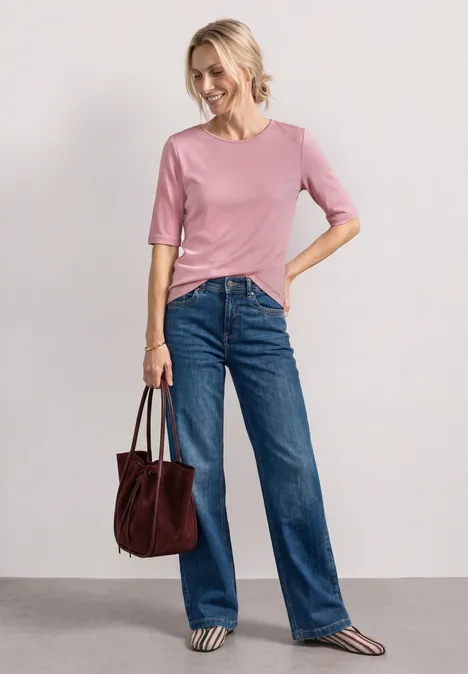 Een vrouw draagt een roze shirt, blauwe spijkerbroek en houdt een bruine handtas vast.