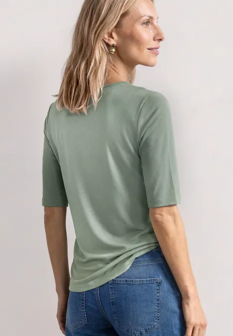 Vrouw van achteren in een saliegroene shirt met halve mouwen en blauwe jeans, blond haar, subtiele oorbel.