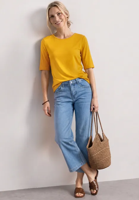Vrouw leunt tegen muur, geel T-shirt, wijde blauwe jeans, gevlochten sandalen en rieten tas.