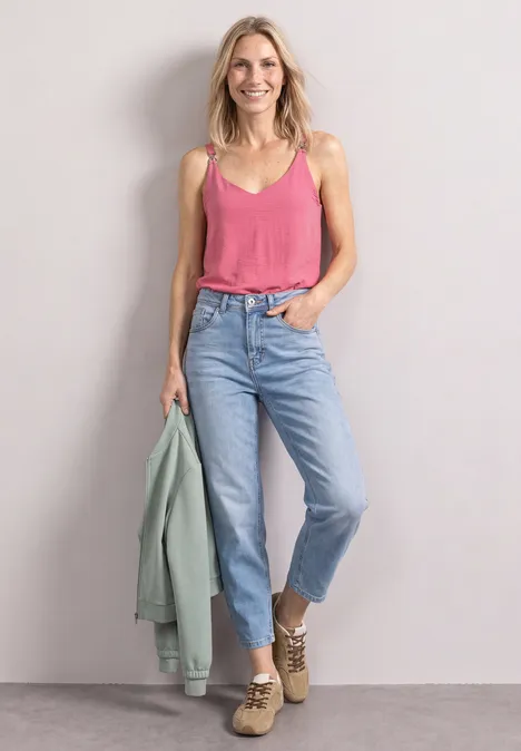 Eine lächelnde Frau in pinkem Top, Jeans, beigen Stiefeletten und grüner Jacke vor grauem Hintergrund.