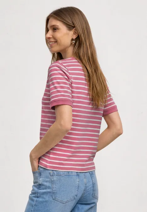 Frau von hinten in gestreiftem rosa-weißem T-Shirt und blauen Jeans, Hände in den Taschen, seitlich abgewandt.