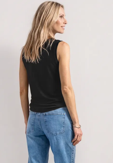Frau von hinten, blonde Haare, schwarzes ärmelloses Top und blaue Jeans vor hellem Hintergrund.