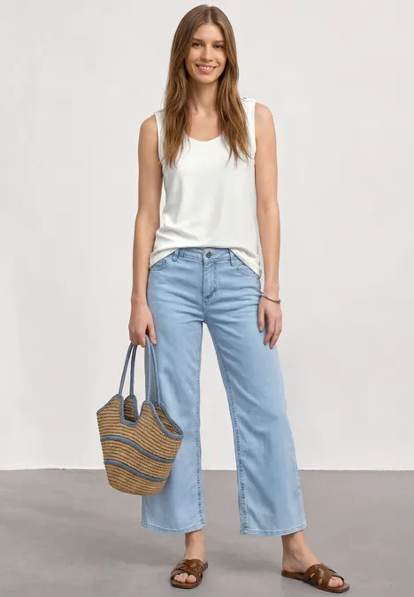 Frau trägt weiße Tanktop, helle Jeans, Sandalen und eine Strohtasche, stehend vor neutralem Hintergrund.
