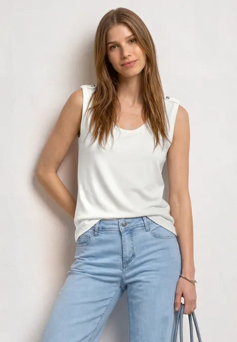 Eine Frau steht vor weißem Hintergrund, trägt ein weißes Top, blaue Jeans und hält ein Handy.