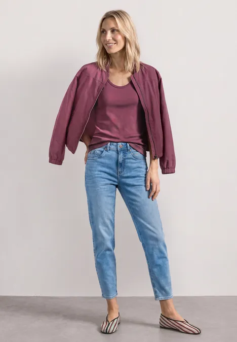 Frau in lila Jacke, dunkelrotem Shirt, hellblauen Jeans und gestreiften Flip-Flops vor neutralem Hintergrund.