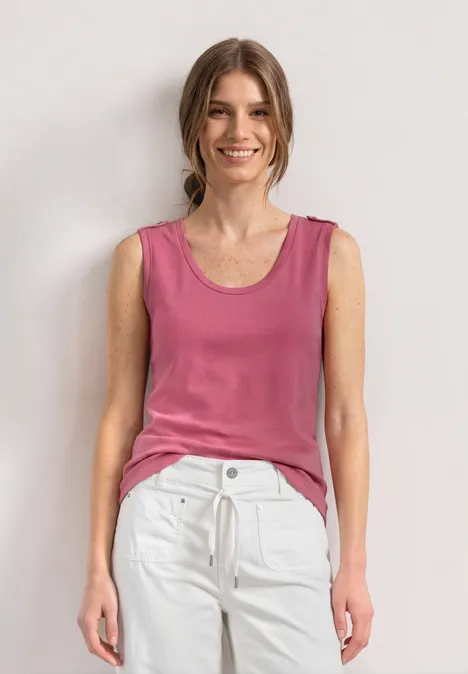 Eine Frau steht vor weißem Hintergrund, trägt rosa Top und weiße Hose.