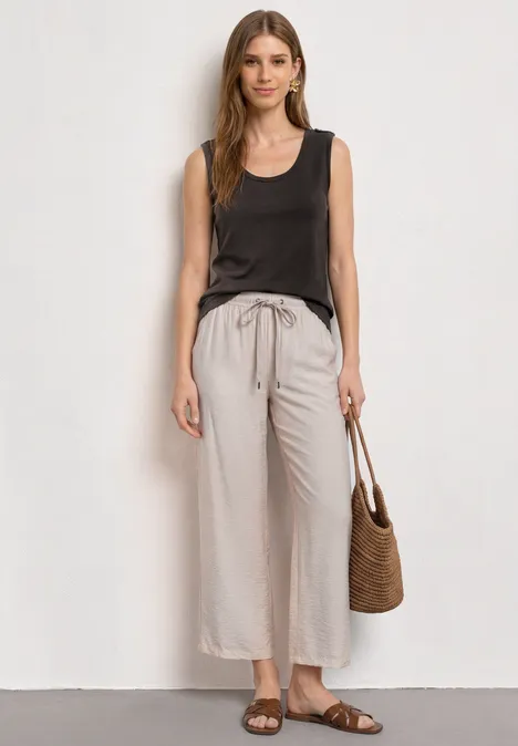 Frau trägt braunes ärmelloses Top, helle, weite Leinenhose, Basttasche und flache Sandalen.