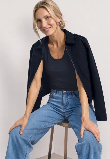 Frau sitzt auf Hocker, trägt blaue Jeans, schwarzes Top und Jacke über den Schultern.