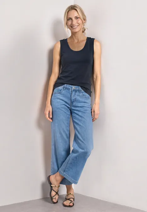 Eine Person in schwarzem Top, weiten Jeans und Sandalen lehnt lässig vor heller Wand.