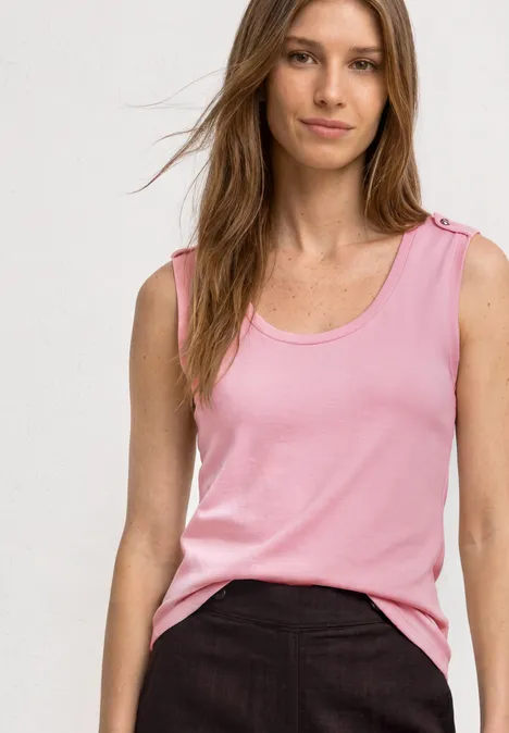Eine Frau trägt ein ärmelloses rosa Top und schwarze Hose vor hellem Hintergrund.