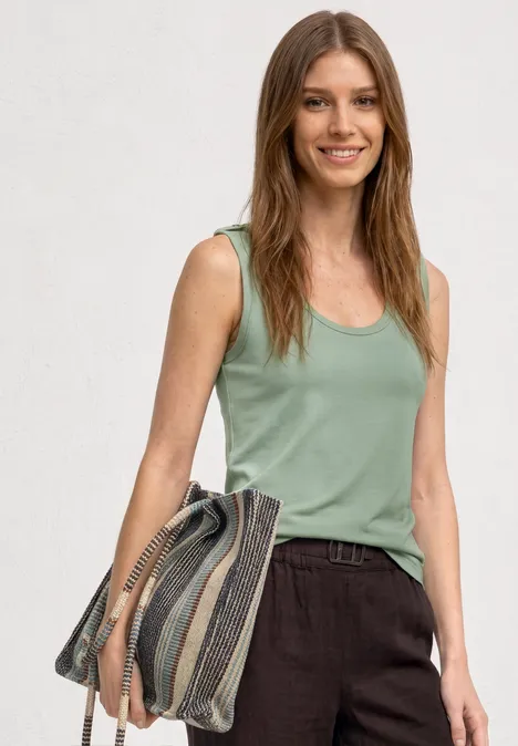 Une femme porte un débardeur vert clair, un pantalon marron et tient un grand sac rayé.