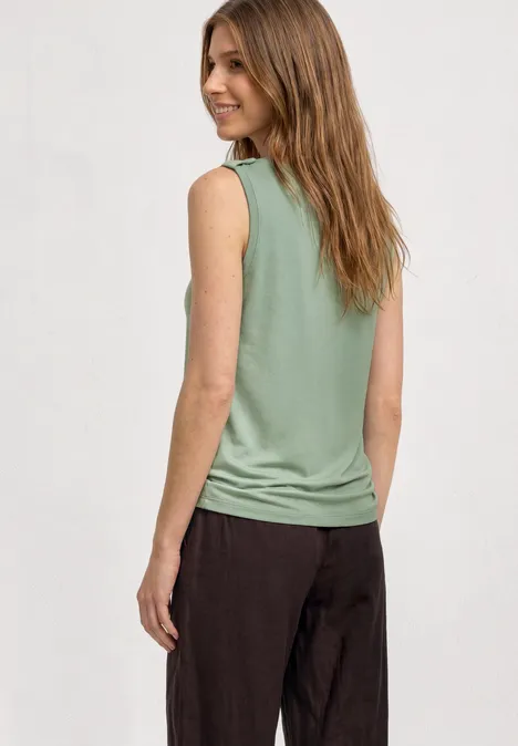 Femme de dos portant un débardeur vert et un pantalon foncé sur fond gris clair.