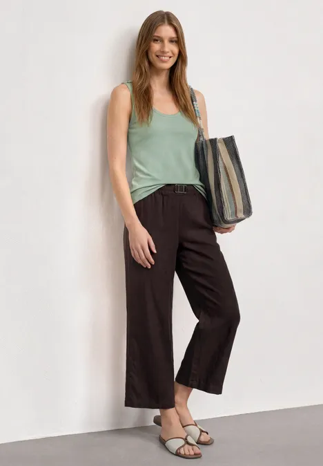 Femme en top vert et pantalon ample foncé, sac à main, sandales sur fond clair.