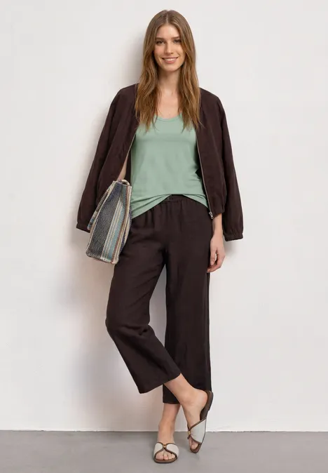 Une femme devant un mur blanc, vêtue d'une veste marron, pantalon ample, top vert menthe, sandales blanches et sac rayé.