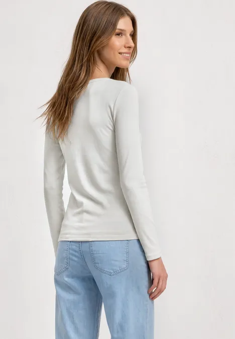Vrouw in wit shirt met lange mouwen en lichtblauwe jeans, kijkt over haar schouder, neutrale achtergrond.
