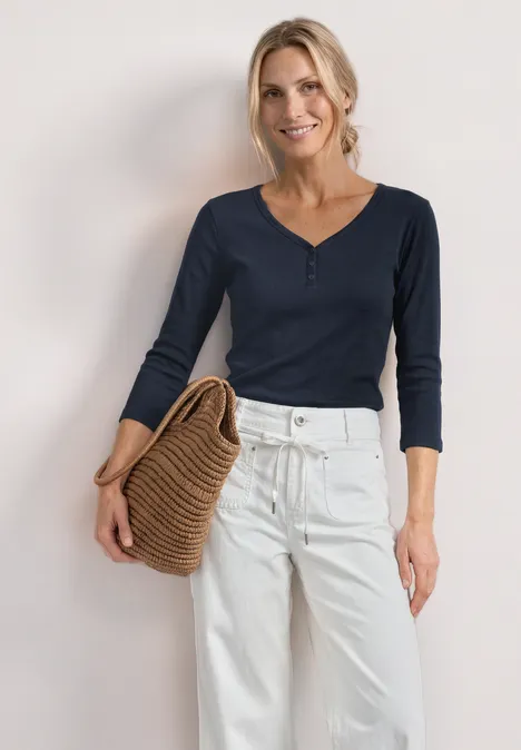 Vrouw in donkerblauwe trui en witte broek, met een strooien tas in haar hand.