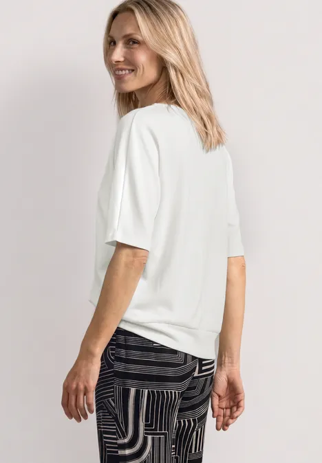 Basic Shirt mit Boat Neck und Elastikbund off white Frau von der Seite mit weißem T-Shirt und schwarz-gemusterter Hose vor hellem Hintergrund.