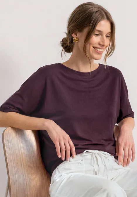 Basic Shirt mit Boat Neck und Elastikbund rustic rouge Eine lächelnde Frau sitzt auf einem Stuhl, trägt ein locker sitzendes lila Oberteil und weiße Hose.