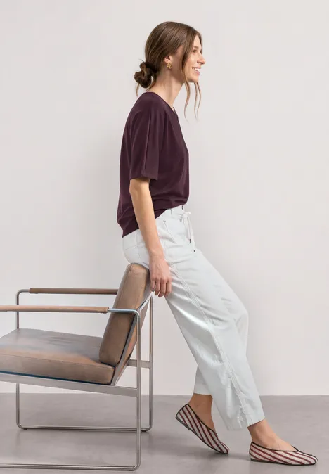 Basic Shirt mit Boat Neck und Elastikbund rustic rouge Frau in violettem T-Shirt, heller Hose und gestreiften Ballerinas lehnt an einem Stuhl vor hellem Hintergrund.