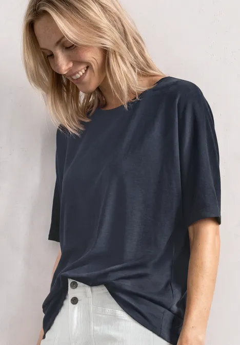 Basic Shirt met boothals en elastische zoom darkest blue Een vrouw in een navyblauw t-shirt en witte broek lacht en kijkt naar beneden tegen een lichte achtergrond.
