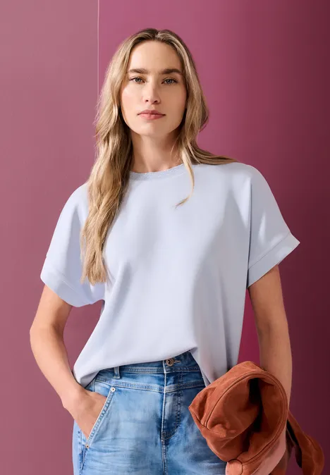 Frau mit hellblauem Shirt hält eine orange Jacke vor rosa Wand