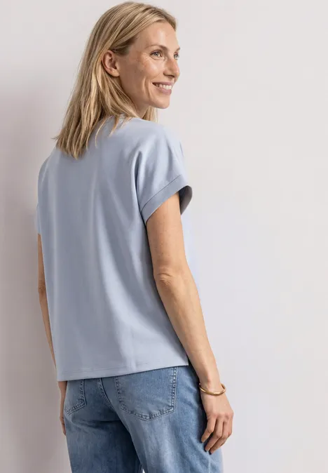 Eine lächelnde Frau in blauem T-Shirt und Jeans, leicht seitlich stehend.