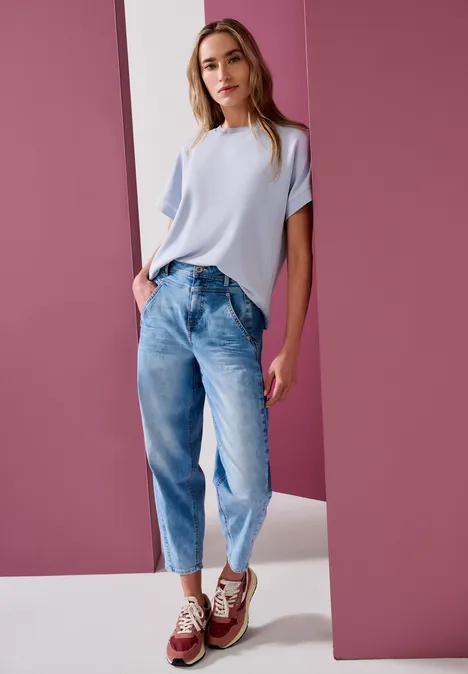 Modell in hellem T-Shirt, weitem Jeans und Sneakers posiert vor rosa Hintergrund.