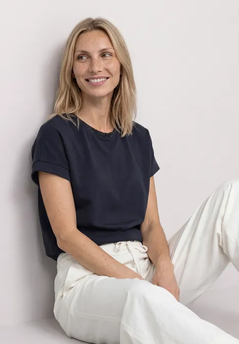 T-shirt avec un aspect soyeux à col rond darkest blue Femme blonde souriante en t-shirt noir et pantalon blanc, assise de manière décontractée devant un mur clair.