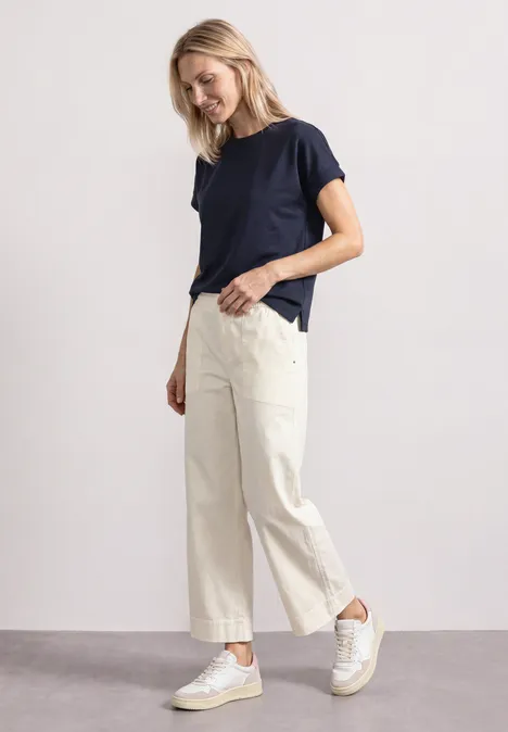 T-shirt avec un aspect soyeux à col rond darkest blue Femme souriante en T-shirt bleu foncé, pantalon crème et baskets blanches, de profil, main dans la poche.