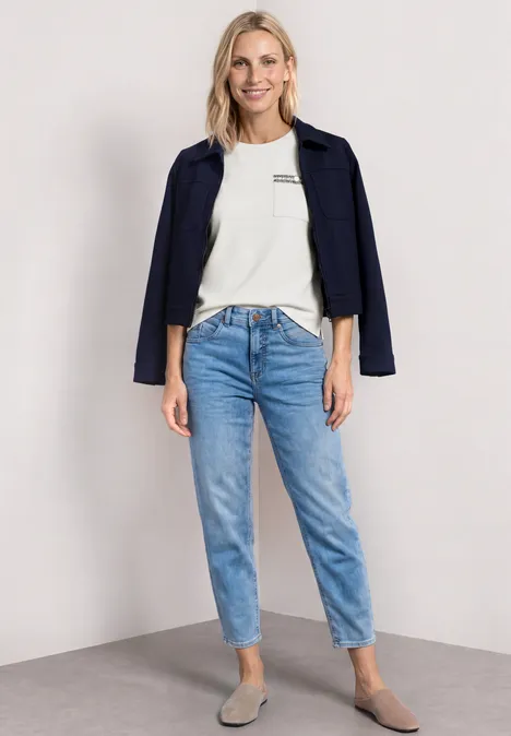 Vrouw met wit T-shirt, donkerblauw jasje op de schouders, lichtblauwe jeans en beige ballerina’s.