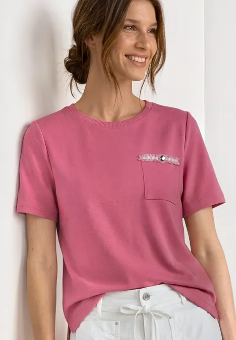 Frau in rosa T-Shirt mit Brusttasche und weißer Hose vor hellem Hintergrund.