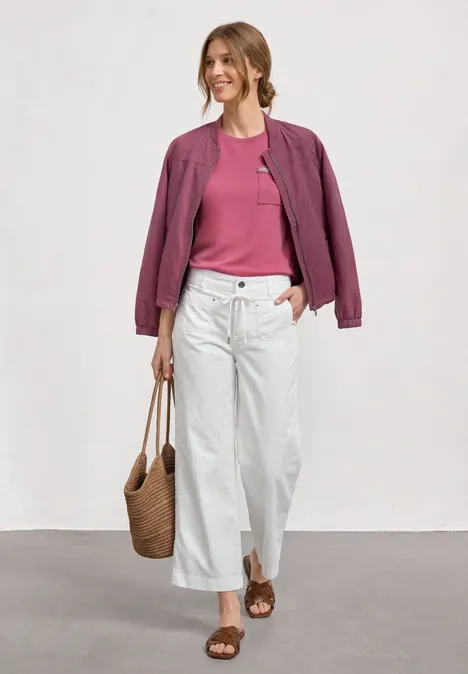 Eine Frau trägt rosa Shirt, violette Jacke, weiße Culotte, Sandalen und Korbtasche beim Gehen.