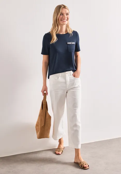 Frau in dunkelblauem T-Shirt, weißen Cropped-Hosen, brauner Tasche und Sandalen vor weißem Hintergrund.
