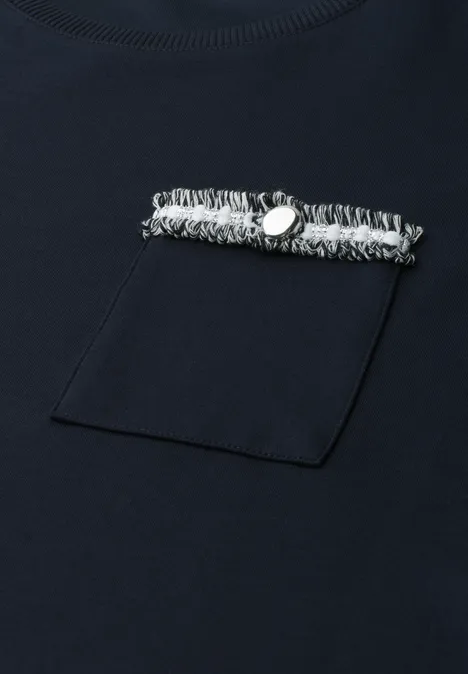 Marineblaues T-Shirt mit Brusttasche und darüber liegender Fransenborte, verziert mit einem Metallknopf.