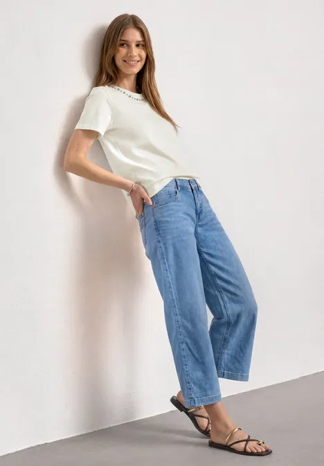 Frau lehnt lässig an Wand, trägt weißes T-Shirt, weite Jeans und schwarze Sandalen.