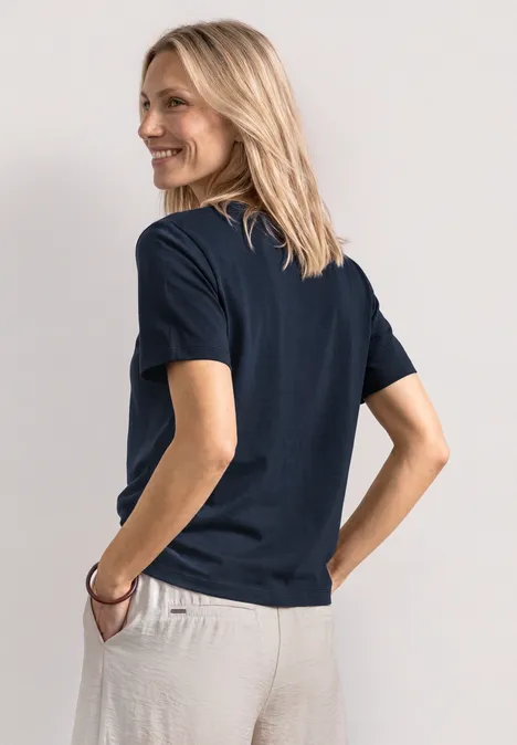 Vue de dos d’une personne portant un t-shirt bleu foncé et un pantalon beige clair, main en poche.