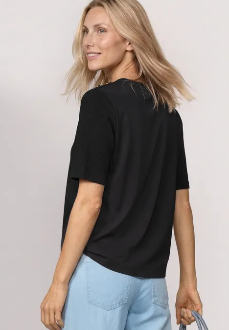 Shirt met ronde hals en knoopdetails Black Vrouw in een zwart T-shirt en blauwe spijkerbroek, schuin van achter gefotografeerd en een tas vasthoudend.