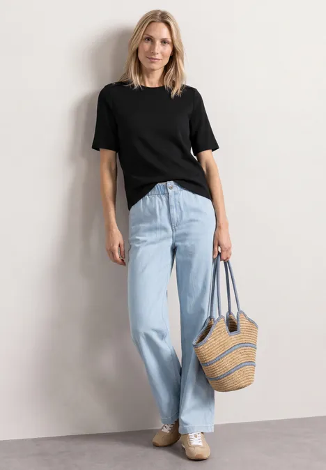 Shirt met ronde hals en knoopdetails Black Vrouw met zwart T-shirt, lichtblauwe jeans en strooien tas voor een witte muur.