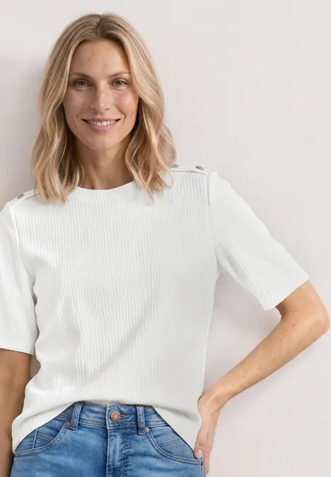 Frau mit schulterlangem blonden Haar trägt ein weißes kurzärmeliges, geripptes Shirt und blaue Jeans.