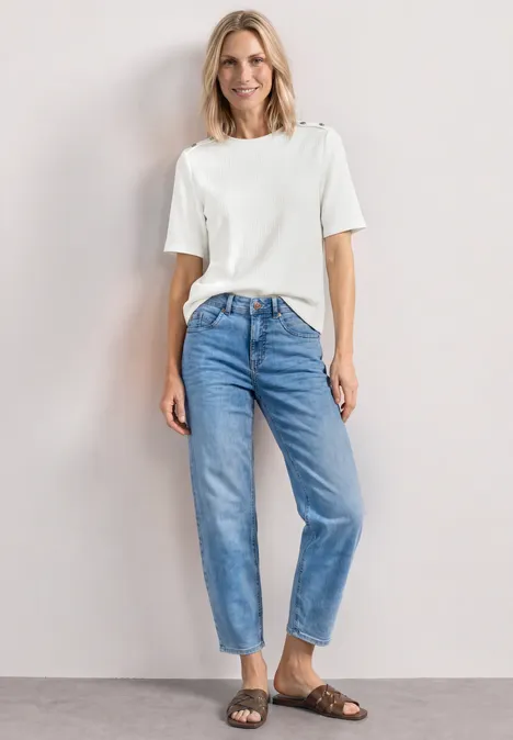Frau trägt weißes T-Shirt, lockere blaue Jeans und braune Sandalen vor hellem Hintergrund.