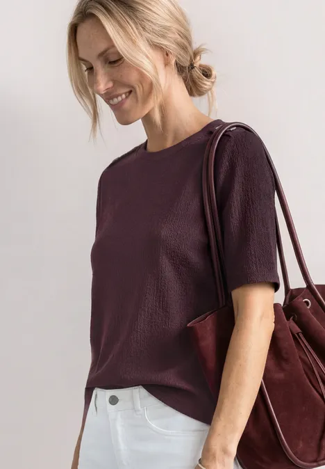 T-Shirt mit Rundhals und Knopfdeatils rustic rouge Frau in einem violetten Kurzarmshirt mit burgunderfarbener Schultertasche vor hellem Hintergrund.