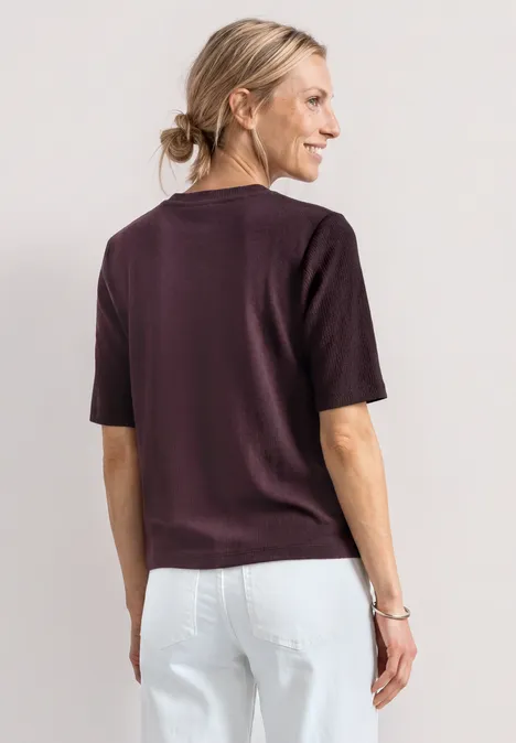 T-Shirt mit Rundhals und Knopfdeatils rustic rouge Frau von hinten in auberginefarbenem T-Shirt und weißer Hose, Haar zu einem Dutt gebunden.