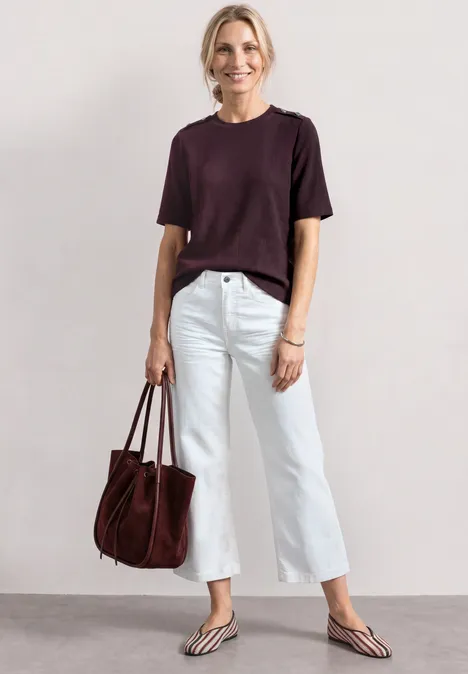 T-Shirt mit Rundhals und Knopfdeatils rustic rouge Person in kurzärmeligem dunkelrotem Shirt, weißen Cropped-Jeans, Flip-Flops und roter Handtasche.