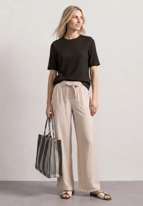 T-Shirt mit Rundhals und Knopfdeatils black coffee Frau in schwarzem T-Shirt und beigen Leinenhosen trägt gestreifte Tasche und Sandalen.