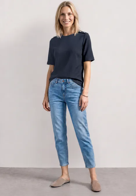 Frau in dunkelblauem T-Shirt, hellblauen Jeans und grauen Slippern vor neutralem Hintergrund.