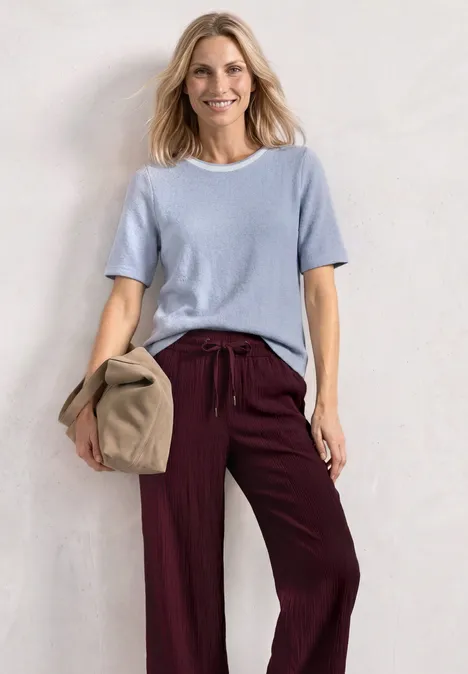 Vrouw met lichtblauw shirt en bordeauxrode broek, houdt een beige tas vast tegen een witte achtergrond.