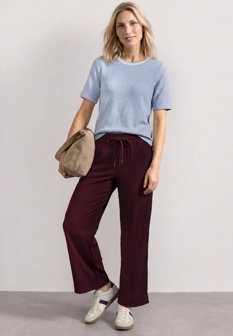 Vrouw in lichtblauw shirt, bordeauxrode broek en witte sneakers, met een hoed in de hand.