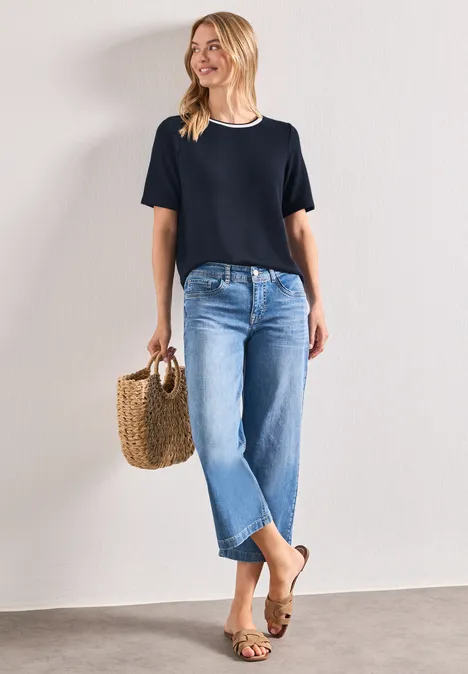 Vrouw leunt glimlachend tegen muur, draagt zwart shirt, blauwe jeans, sandalen en een gevlochten mand.