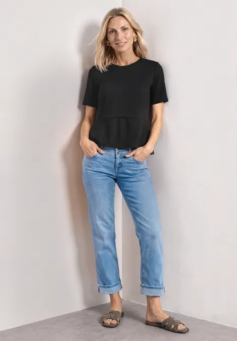 T-Shirt mit Rundhals und Volant-Detail Black Frau in schwarzem T-Shirt, blauen Jeans und Sandalen steht vor hellem Hintergrund.